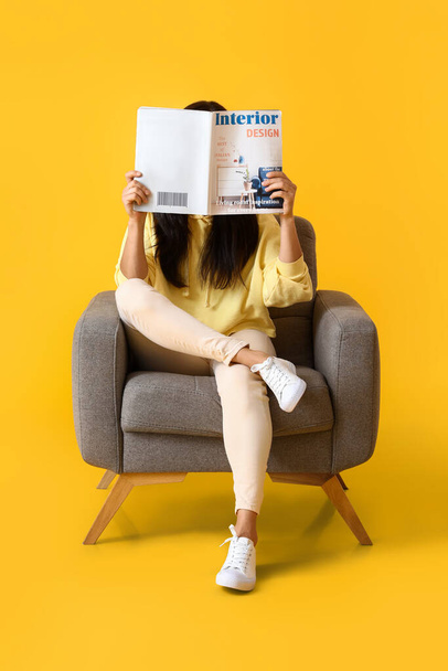 Beautiful young woman reading magazine in soft armchair on yellow background - Photo, image
