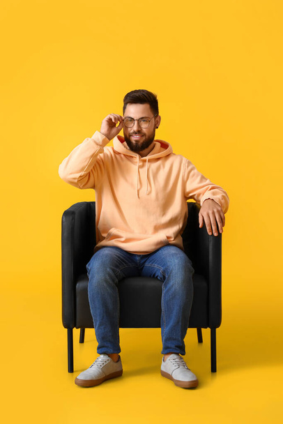 Handsome bearded man with eyeglasses sitting in leather armchair on yellow background - Фото, зображення