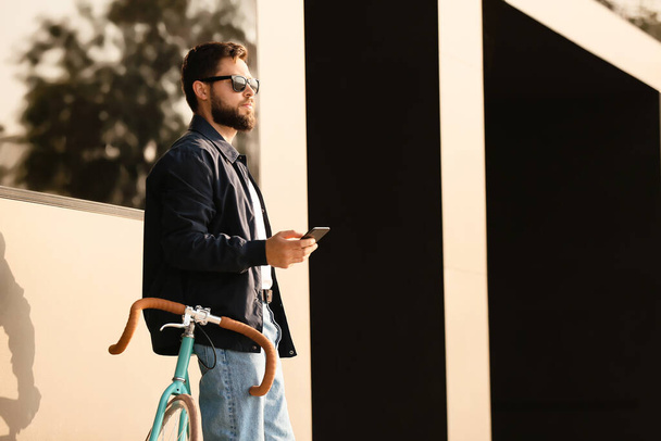 Elegante hombre barbudo con bicicleta usando el teléfono móvil en la ciudad - Foto, imagen