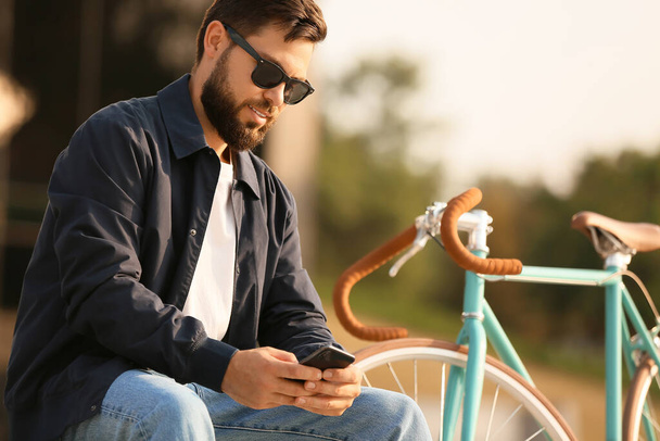 Stijlvolle man met baard en fiets met mobiele telefoon in de stad - Foto, afbeelding