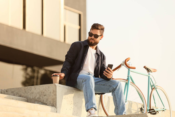 Stílusos szakállas férfi mobiltelefonnal, csésze kávé és kerékpár a városban - Fotó, kép