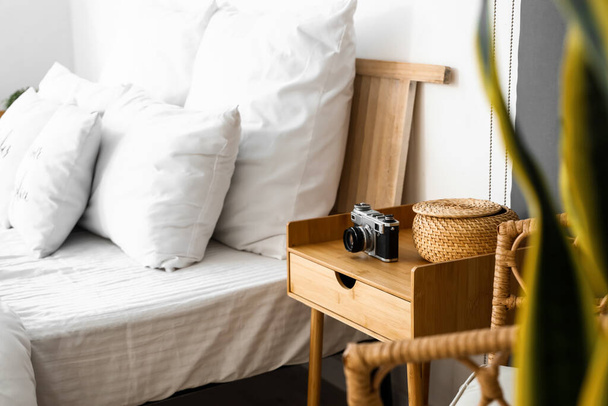 Mesita de noche de madera con cámara fotográfica y cesta de mimbre en dormitorio luminoso - Foto, Imagen