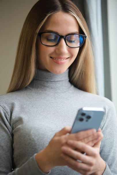 Happy blonde woman in glasses typing message online on modern mobile phone connected to internet. Cheerful white female using new smartphone for online communication and entertainment - Fotografie, Obrázek