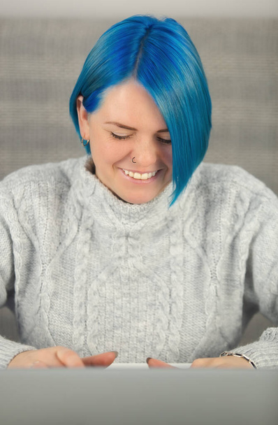 Heureuse femme blanche aux cheveux bleus qui travaille sur un ordinateur portable avec un sourire joyeux. Jolie jeune femme adulte faisant du travail à distance à la maison en confinement. Entrepreneur femme d'affaires tapant sur le clavier  - Photo, image