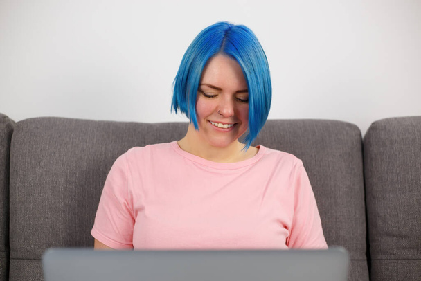 Happy freelancer woman with blue hair working on notebook computer at home. Cheerful white female with dyed bob haircut wearing casual pink t-shirt doing distant work online on lockdown - Photo, Image