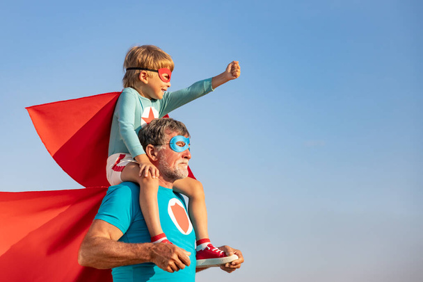 Superhero senior man and child playing outdoor. Super hero grandfather and boy having fun together against blue summer sky background. Family holiday concept. Happy Father's day - 写真・画像