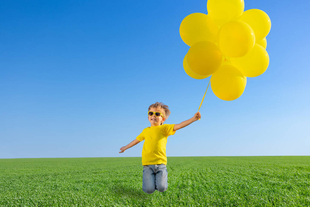 Happy child jumping in green spring field. Kid with yellow balloons against blue sky background. Carefree and freedom outdoor - Zdjęcie, obraz