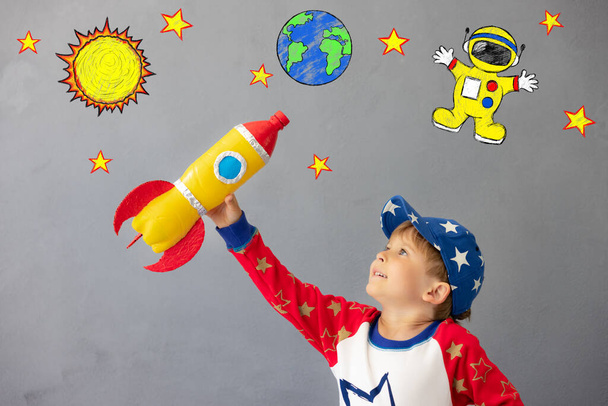 Happy child playing with toy rocket against concrete wall background. Kid pretend to be astronaut. Imagination and children dream concept - Zdjęcie, obraz