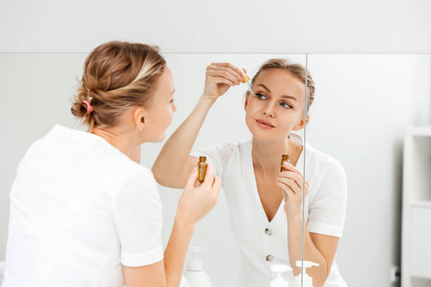 A young woman looks in the mirror and applies cosmetic oil on her face. Back view from the shoulder. Skin care concept. - Photo, Image