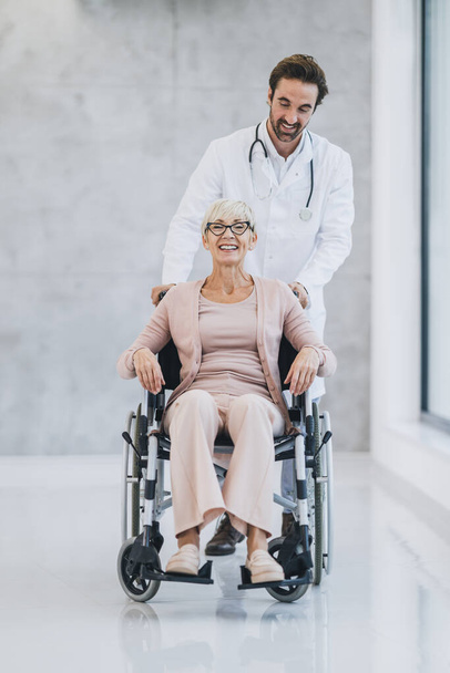Girato di un giovane medico che spinge la sua paziente anziana sulla sedia a rotelle e parla con lei. - Foto, immagini