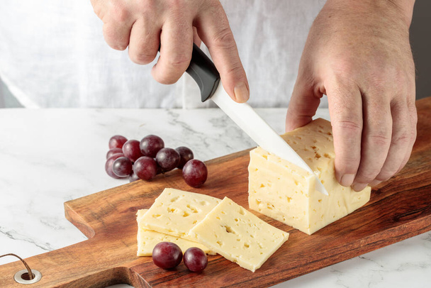 男性の手は、キッチンの木製のまな板にセラミックナイフでハードチーズをカットします。コピースペースの大理石の背景 - 写真・画像