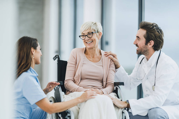 Médico de sucesso e jovem enfermeira conversando com sua paciente idosa cadeirante no corredor do hospital. - Foto, Imagem