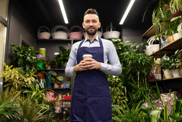 jardinero en delantales y una taza de café en las manos en el fondo de una floristería - Foto, Imagen