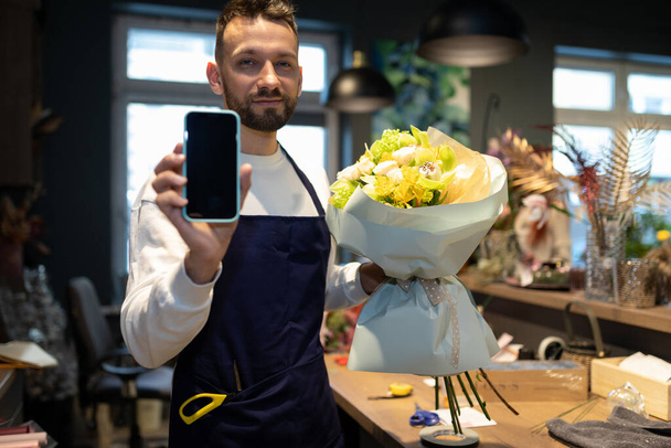 флорист с букетом в руках демонстрирует экран смартфона на фоне цветочного магазина - Фото, изображение
