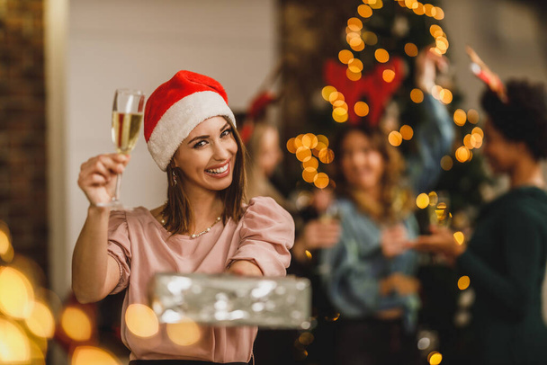 gelukkig jong vrouw brengen geschenken en toasten met champagne als ze vieren Kerstmis met haar vrienden thuis feest. - Foto, afbeelding