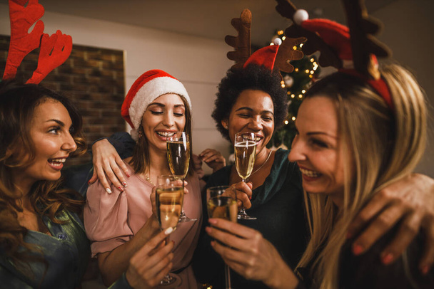 Четыре весёлых многонациональных девушки пьют за шампанское на Рождество или новогоднюю домашнюю вечеринку. - Фото, изображение