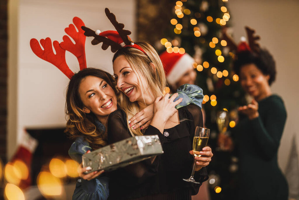 Glimlachende vriendinnen knuffelen en geven cadeautjes aan elkaar tijdens het verzamelen thuis om Kerstmis of Nieuwjaar te vieren. - Foto, afbeelding