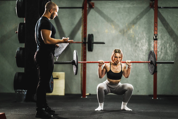 Personal trainer watching and assisting a muscular young woman in weight training at the gym. She is doing squat exercise with barbell. - 写真・画像