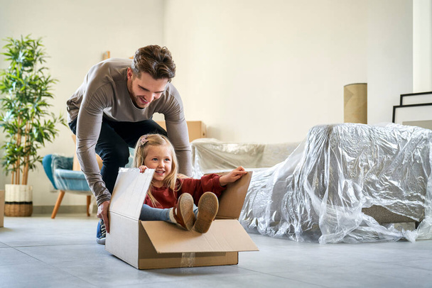 Caucasian father pushing little girl in paper box while moving house  - 写真・画像