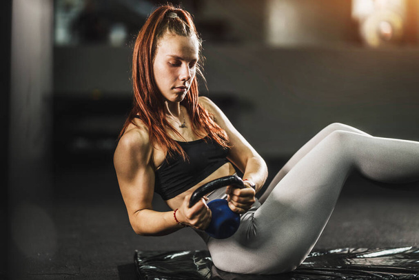 Zastřelen svalnaté mladé ženy ve sportovním oblečení dělat sedí s kettlebell při cvičení v tělocvičně. - Fotografie, Obrázek
