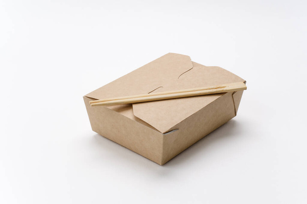 Baguettes sur une boîte fermée en carton pour la livraison de nourriture sur un fond blanc. Plats rectangulaires écologiques pour la livraison de la cuisine asiatique. Modèle de publicité. - Photo, image