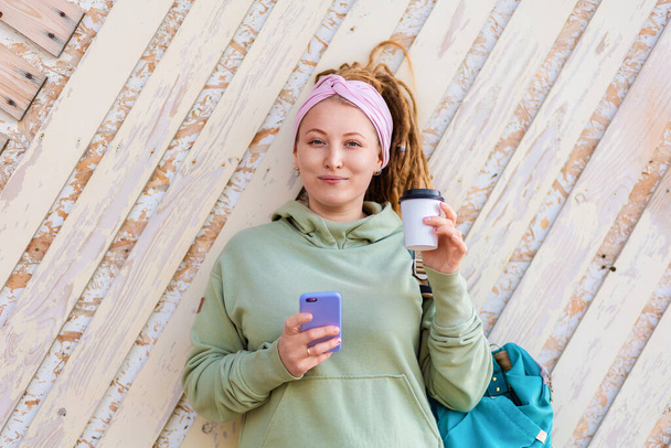 Portrait of beautiful young woman with dreadlocks poses against a pink wooden wall with a cup of coffee, a phone and a backpack. The concept of business and freelancing. - Фото, зображення