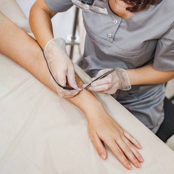 A female cosmetologist performs the procedure of permanent removal of unwanted hair in the forearm by electroepilation device and tweezers. - Foto, Bild