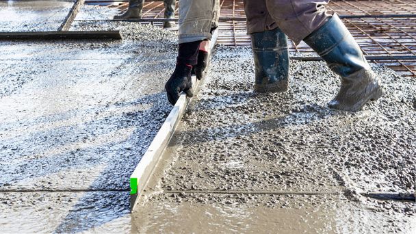 Építőipari munkás szintező öntött beton padló egy ipari műhelyben. Lábak csizmában a betonban. Felszíni betonozás. Monolitikus vasbeton gyártása - Fotó, kép