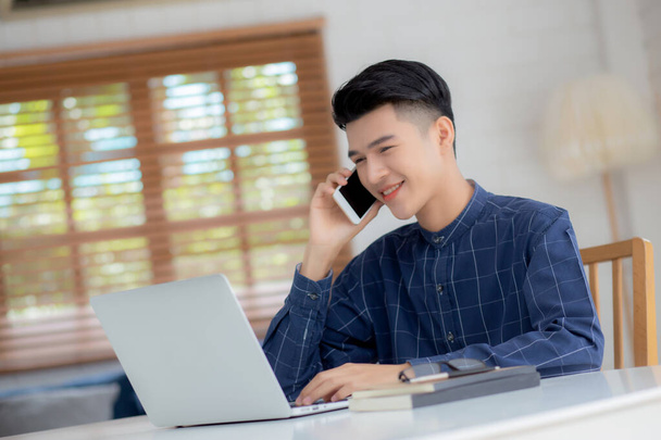 Mladý asijský muž mluví telefon a práci z domova s notebookem, na volné noze pomocí notebooku na internet on-line a mluvit na smartphone, domácí život, obchodní a komunikační koncept. - Fotografie, Obrázek