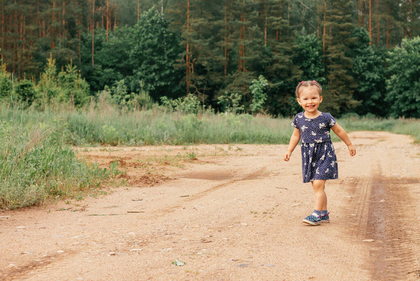 Щаслива маленька дівчинка 2-3 роки в блакитній сукні і сандалі біжить по сільській дорозі в літньому полі біля лісу
 - Фото, зображення