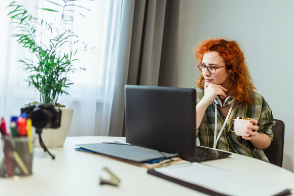 Zdalne życie zawodowe. Młoda rudowłosa kobieta pracuje na laptopie w domu. - Zdjęcie, obraz