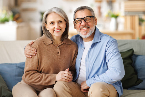 Porträt eines glücklichen Senioren-Familienpaares, das sich umarmt, während es zu Hause auf dem Sofa sitzt - Foto, Bild