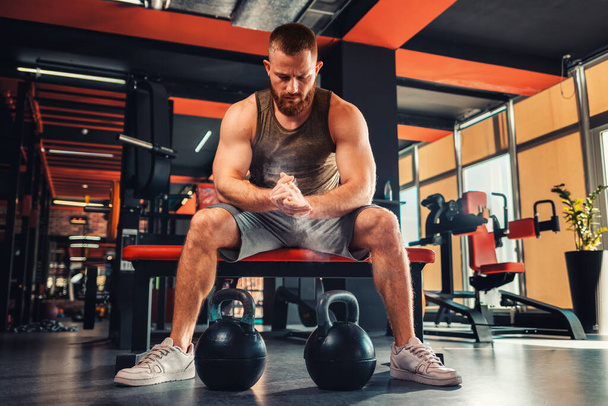 A young bearded athletic man trains in the gym with dumbbells. The athlete rubs magnesia in his hands. The concept of fitness. - Foto, afbeelding