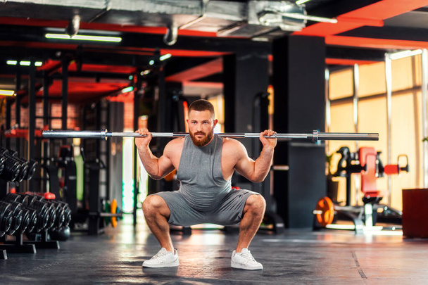 A young bearded athletic man trains in the gym squatting with a barbell. The concept of fitness and training. - Foto, Imagen