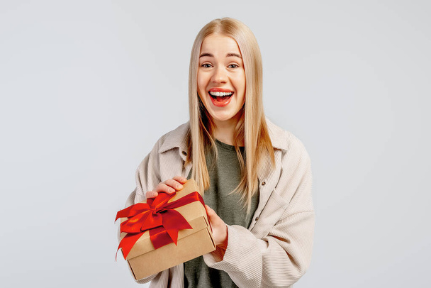 Sorrindo jovem loira segurar caixa de presente com fita vermelha, de pé isolado no fundo claro. Ano Novo Conceito de feriado de aniversário do Dia das Mulheres - Foto, Imagem