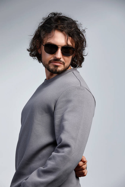 Retrato de moda de hombre elegante guapo con pelo rizado usando suéter gris y gafas sobre fondo gris en el estudio - Foto, Imagen