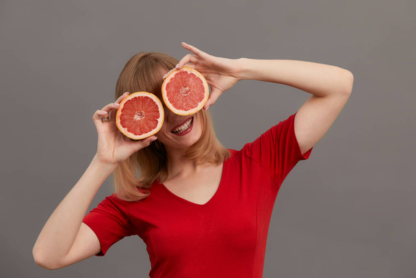 Beautiful woman with citrus fruits in hands at the eyes. Model with long hair hold cut grapefruit in hands as glasses isolated on gray. Skin care concept. Healthcare concept. - Fotó, kép