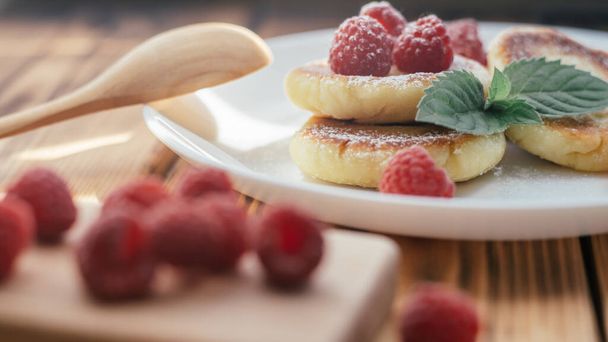 Heerlijke cheesecakes of pannenkoeken met poedersuiker op wit bord op houten tafel met frambozen en muntblad - Foto, afbeelding