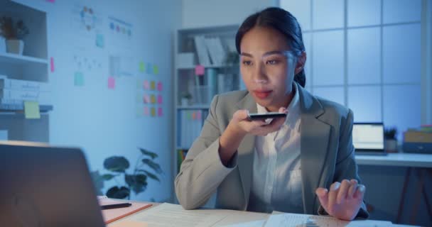 Νεαρή Ασία χαρούμενα επαγγελματική γυναίκα κάθεται στο γραφείο με φορητό υπολογιστή ηχογράφηση ηχητικό μήνυμα στο smartphone επιβεβαιώσει λεπτομέρεια στο γραφείο τη νύχτα. Γυναικείο κοστούμι, Τεχνολογία startup επιχειρηματική ιδέα. - Πλάνα, βίντεο