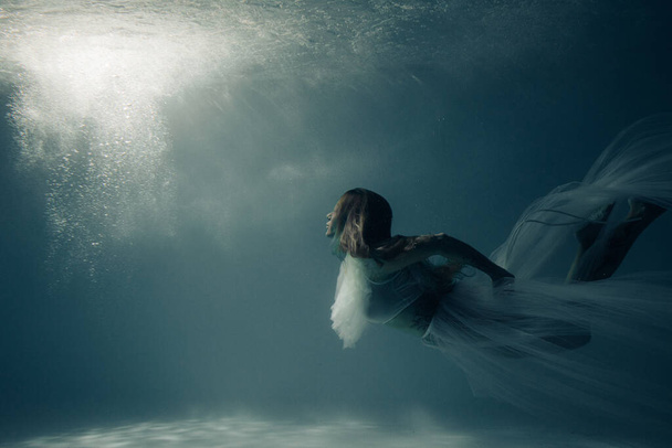 Όμορφη γυναίκα με νυφικό φόρεμα κολυμπά και καταδύεται κάτω από ηλιόλουστα βαθιά νερά. - Φωτογραφία, εικόνα