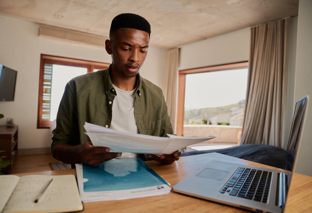 Focalisé jeune adulte noir mâle étudiant avec des notes en papier, livre et ordinateur portable dans un appartement moderne.  - Photo, image