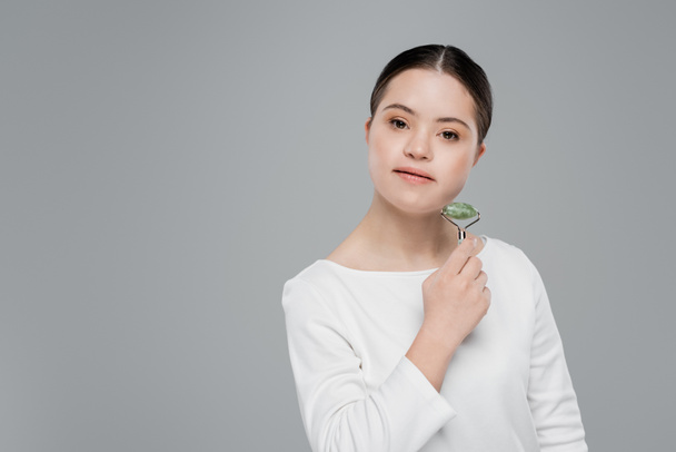 Young woman with down syndrome holding jade roller isolated on grey  - Φωτογραφία, εικόνα