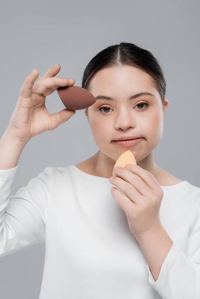 Woman with down syndrome holding beauty blenders isolated on grey  - Foto, imagen