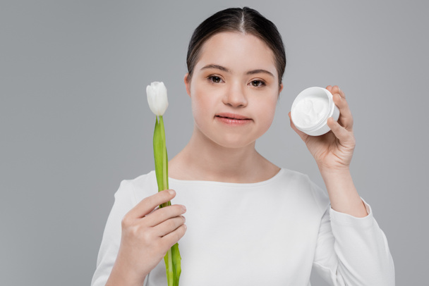 Brunette woman with down syndrome holding tulip and cosmetic cream isolated on grey  - Fotó, kép
