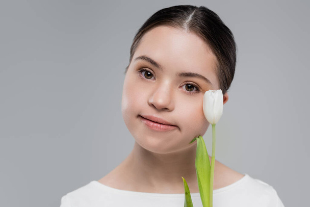 Young woman with down syndrome holding tulip near face isolated on grey  - Foto, afbeelding