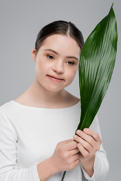 Smiling brunette woman with down syndrome holding leaf isolated on grey  - Foto, Imagem