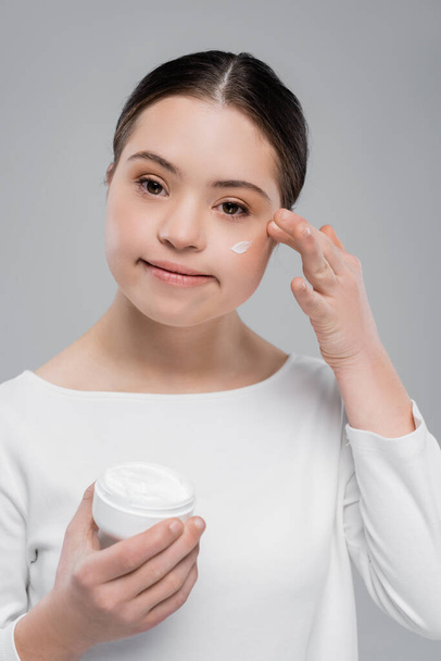 Mujer joven con síndrome de Down aplicando crema facial aislada en gris  - Foto, imagen