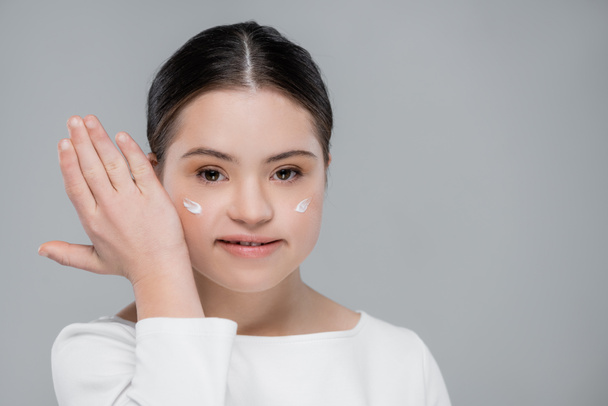 Positive woman with down syndrome and face cream isolated on grey  - Zdjęcie, obraz