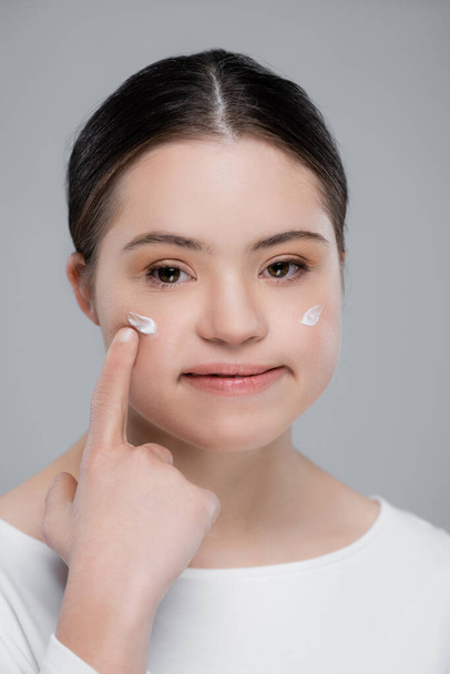 Woman with down syndrome pointing at cream on face isolated on grey  - Φωτογραφία, εικόνα