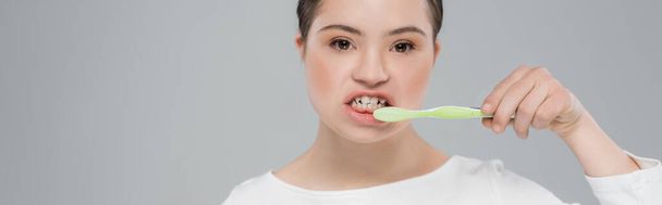 Νεαρή γυναίκα με σύνδρομο Down βουρτσίζει τα δόντια απομονώνονται σε γκρι, πανό  - Φωτογραφία, εικόνα